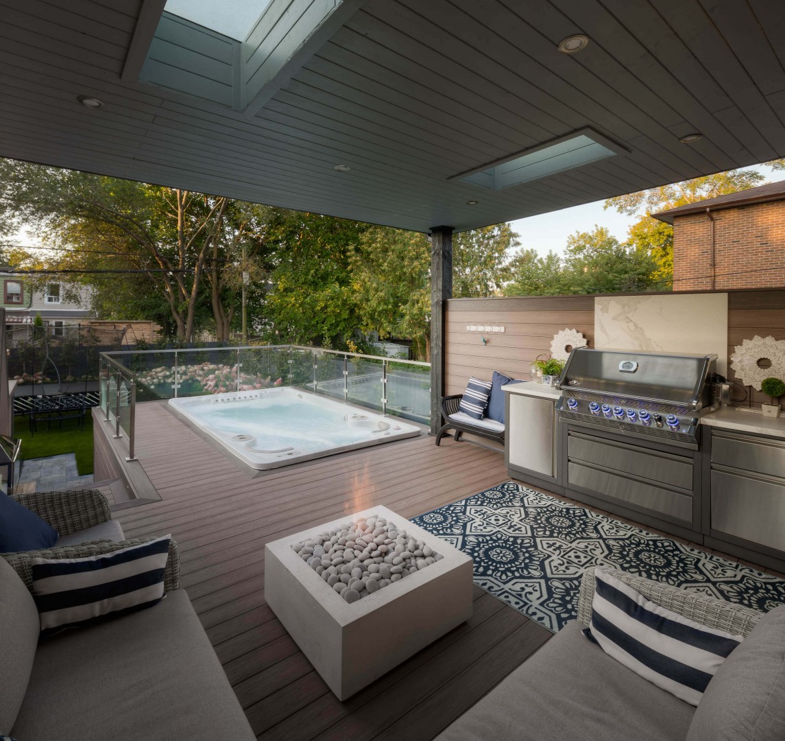 An image of an outdoor covered deck, featuring a hot tub, stainless-steel barbeque and outdoor conversation set.