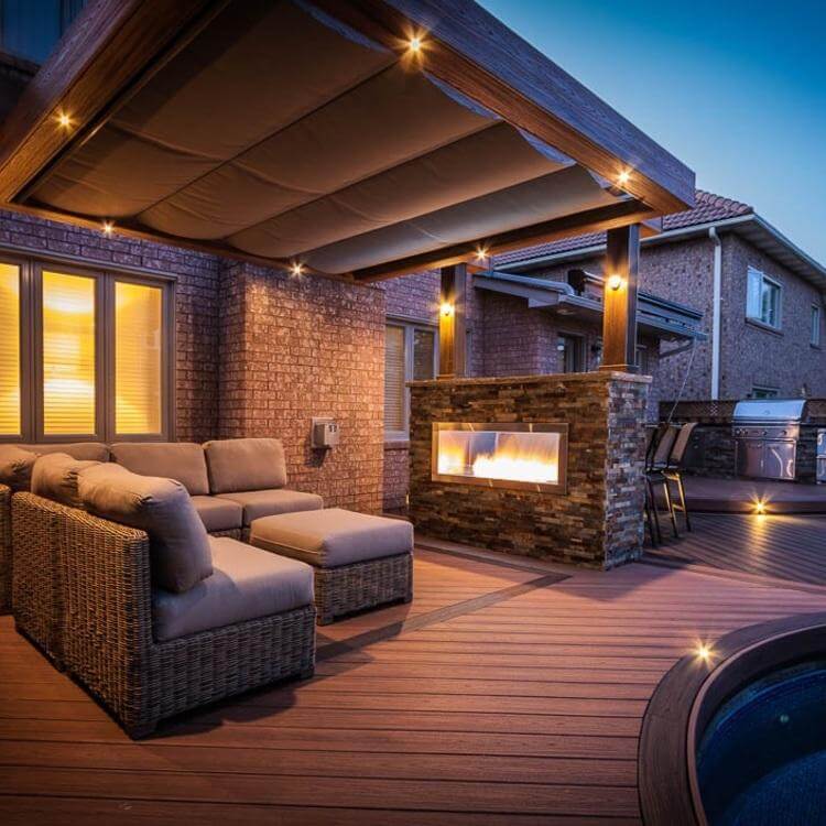 A photo of a covered deck at dusk, lit up by overhead pot lights and an outdoor stone fireplace. There is a large beige sectional, and a built-in pool.