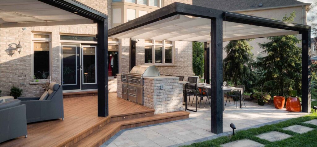 Deck with stone work breaking up the space, an outdoor kitchen for gathering, and pergolas above for shade