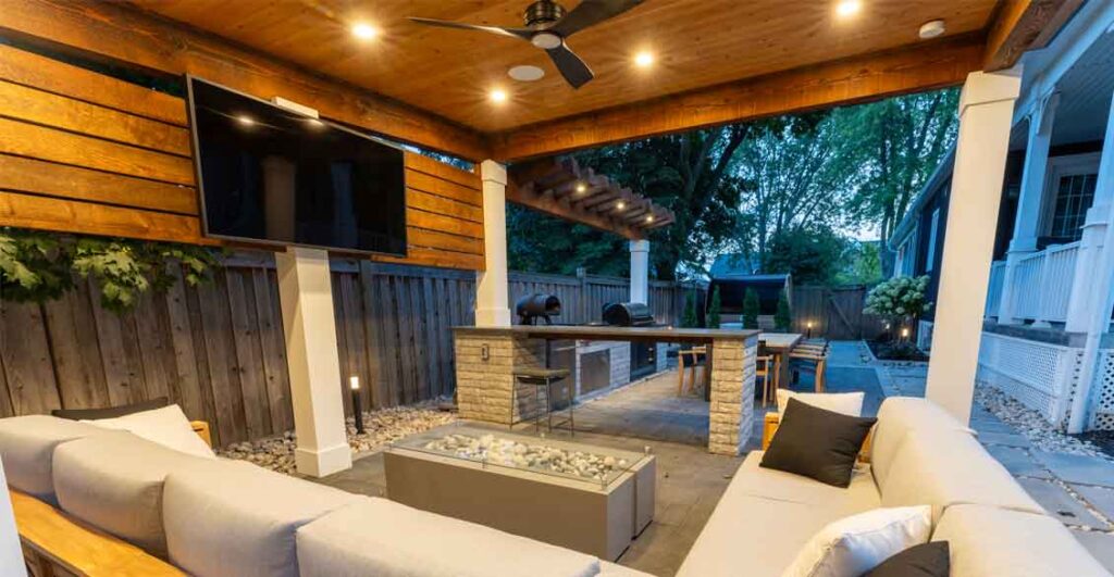Outdoor cabana with sofa and TV, creating a backyard living room beside a dining area and outdoor kitchen