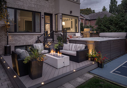 Two-level Wolf PVC deck in Toronto with upper swim spa access, covered BBQ area, lower fireplace lounge, and evening lighting surrounded by mature gardens.