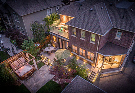 Resort-style Oakville backyard with a glass-rail deck, lush garden beds, stone patio, and cedar pergola over a hot tub with string lights.